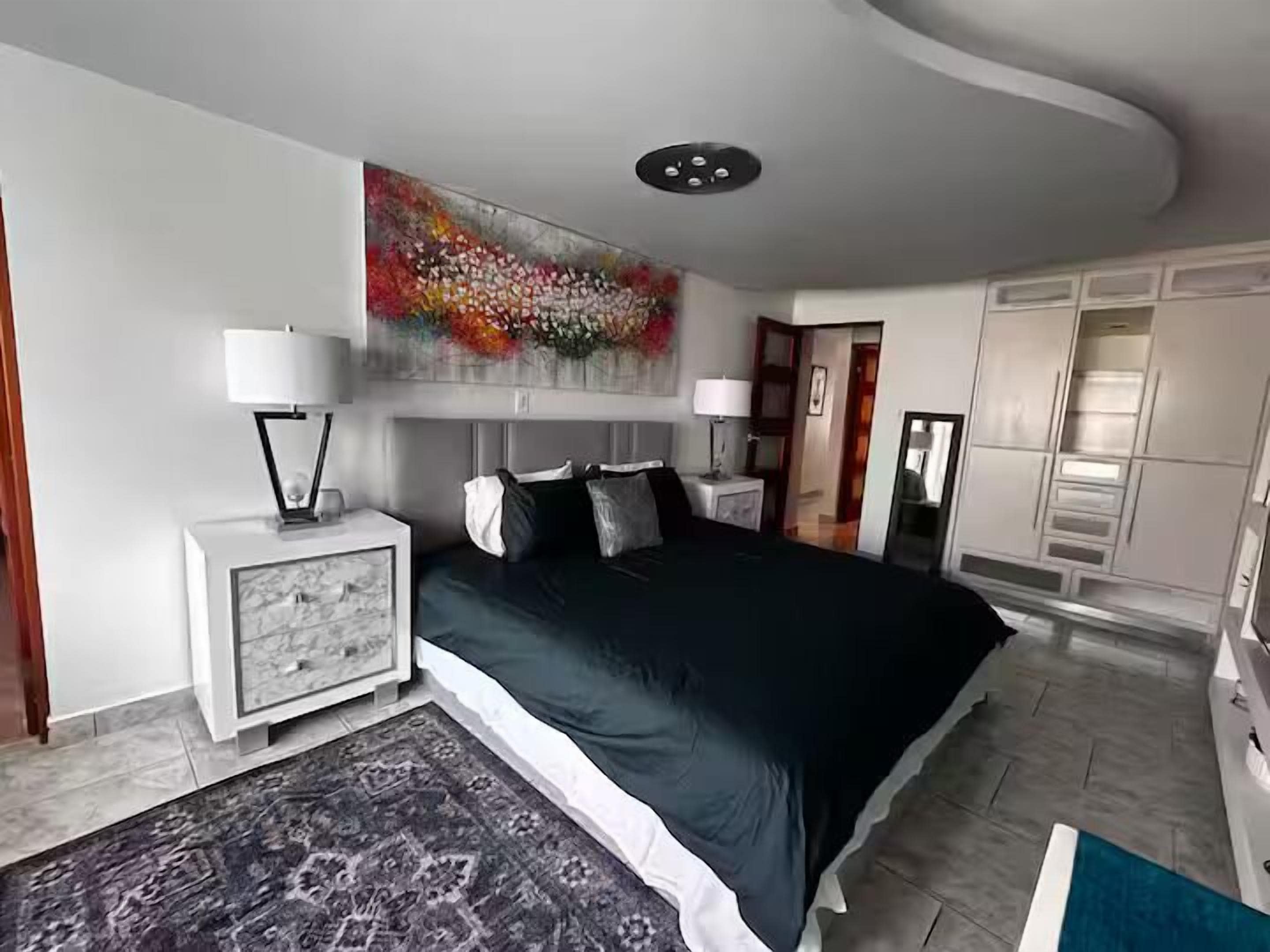A modern bedroom with a large bed, a nightstand, a side table, a decorative wall piece, and a wardrobe.