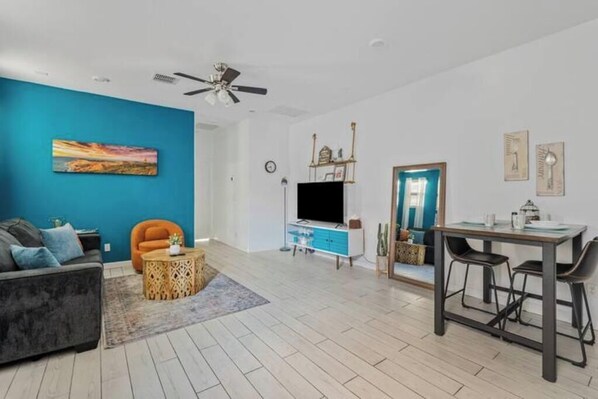 A modern living room with a blue accent wall, a sofa, a coffee table, a TV, a dining table with chairs, and a ceiling fan.
