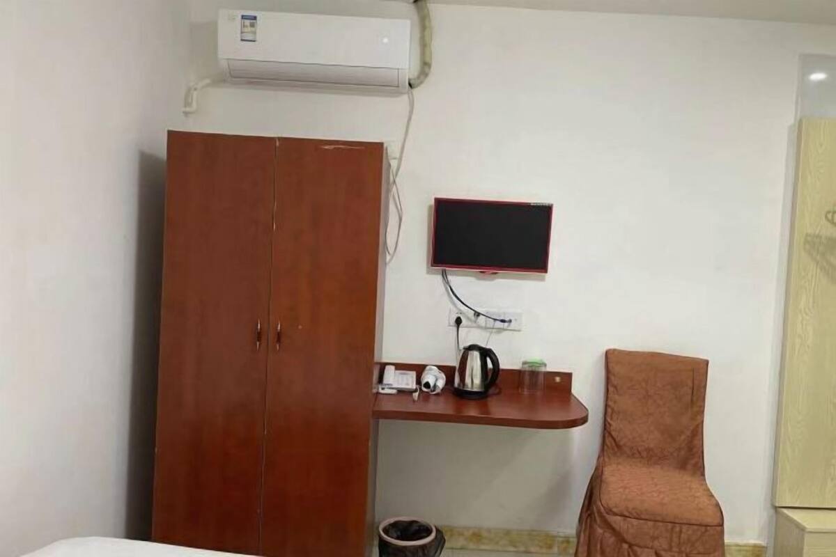 A room with a wardrobe, a desk with a computer monitor, a chair, and a trash bin.