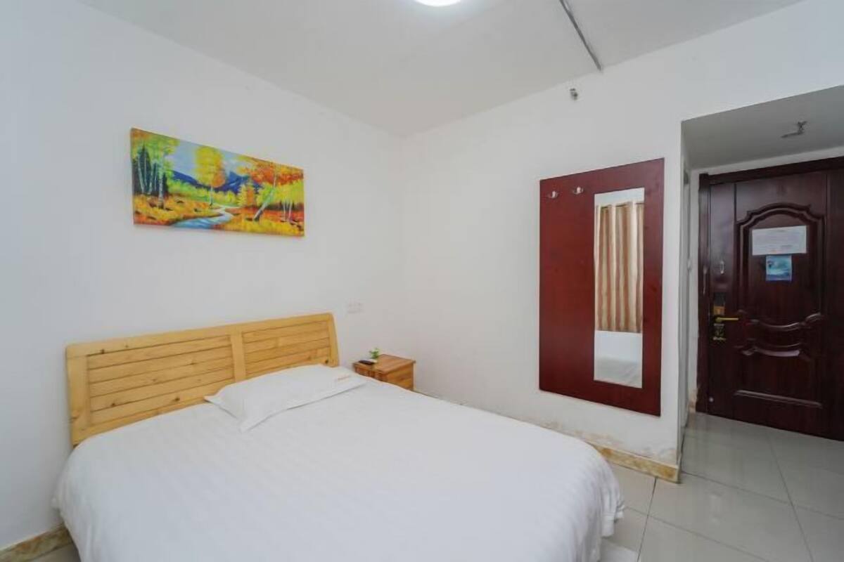 A room with a wooden bed, a small bedside table, a colorful painting on the wall, and a red door.