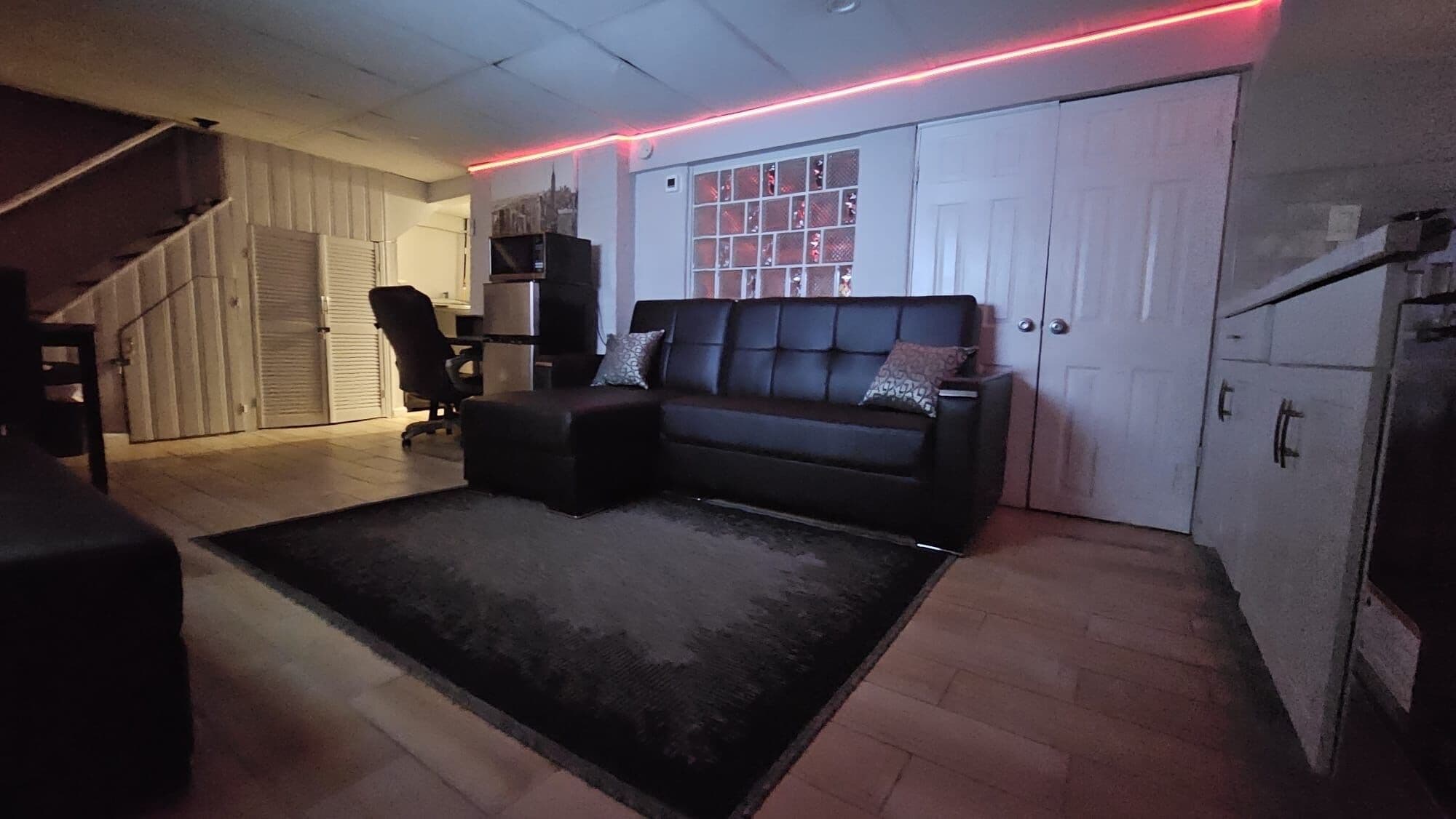 A living room with a black leather sofa, a black armchair, and a patterned rug. There is a staircase leading to another level, a door with a window, and a white cabinet.