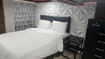 A neatly made bed with white linens, a headboard with a geometric pattern, and a small bedside table with drawers.