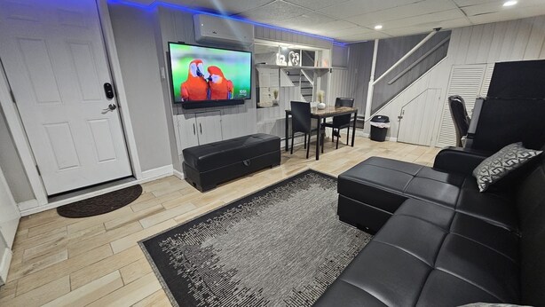 A modern living room with a black leather sofa, a flat-screen TV, a dining area with a table and chairs, and a staircase. - Lofty Cozy Suite near Nassau Coliseum and Hofstra University (Hempstead)
