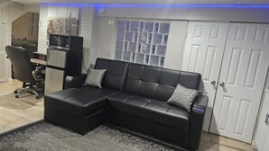 A modern living room with a sectional sofa, a black office chair, a small TV, and a decorative window with a grid pattern. - Lofty Cozy Suite near Nassau Coliseum and Hofstra University (Hempstead)