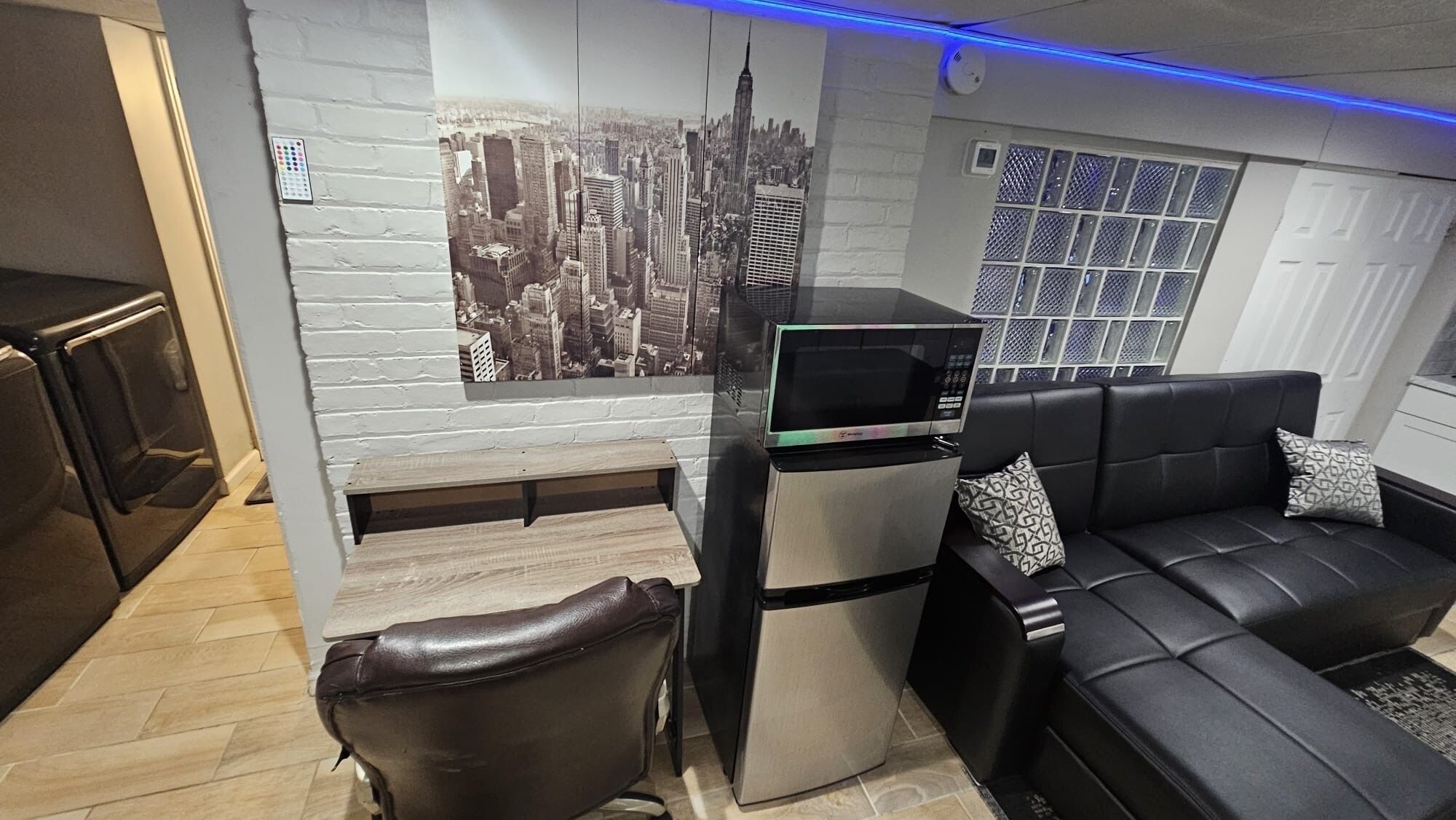 Ein kompakter Wohnraum mit einer Küchenzeile, einem schwarzen Ledersofa, einer Holzbank und einer Wandmalerei mit Stadtansicht.