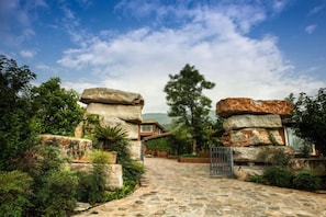 A stone pathway leading to a building with a metal gate, surrounded by greenery and large rock formations.