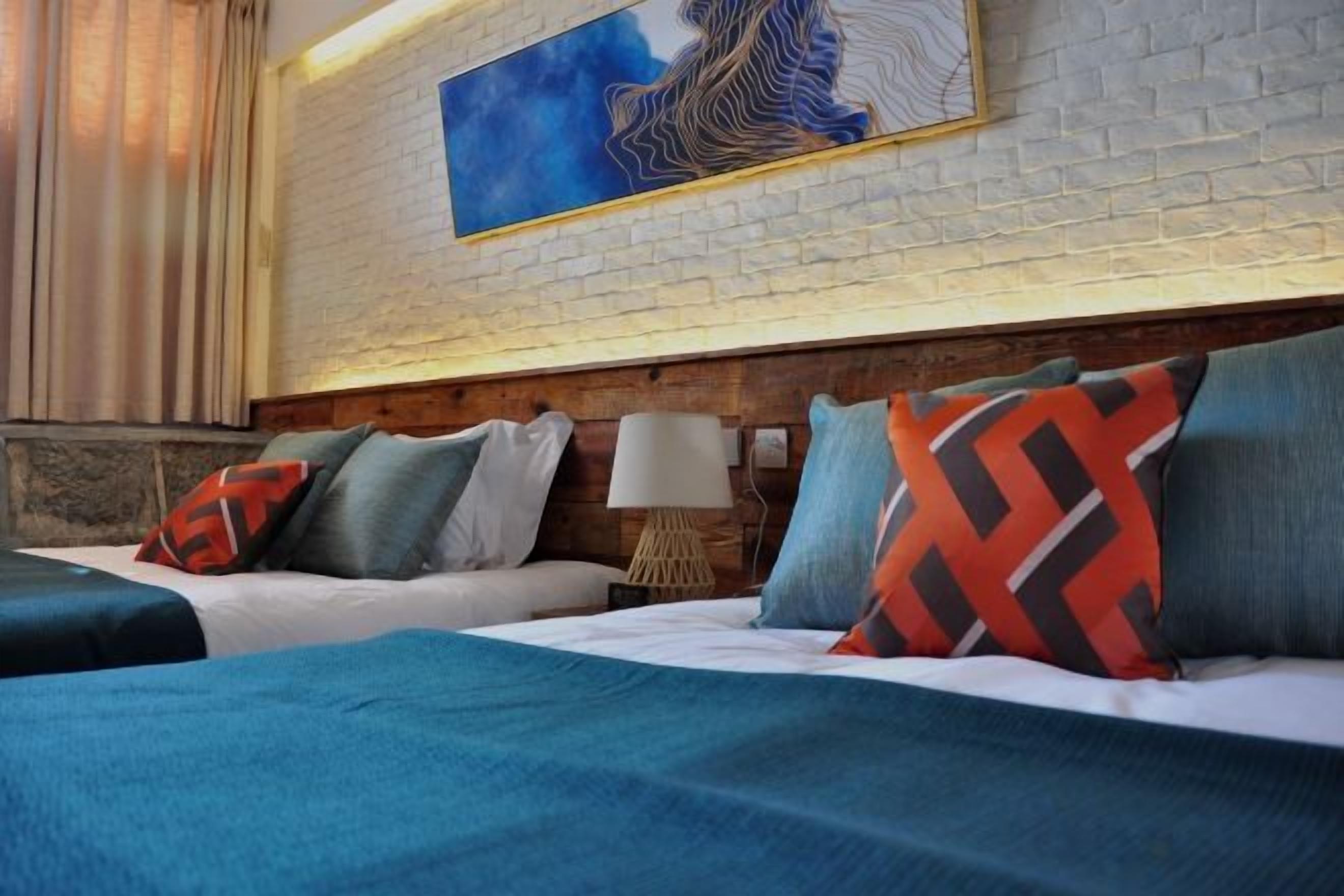 Una camera d'albergo con parete in mattoni, un quadro di grandi dimensioni, due letti con coperte dai colori differenti e un comodino con una lampada.
