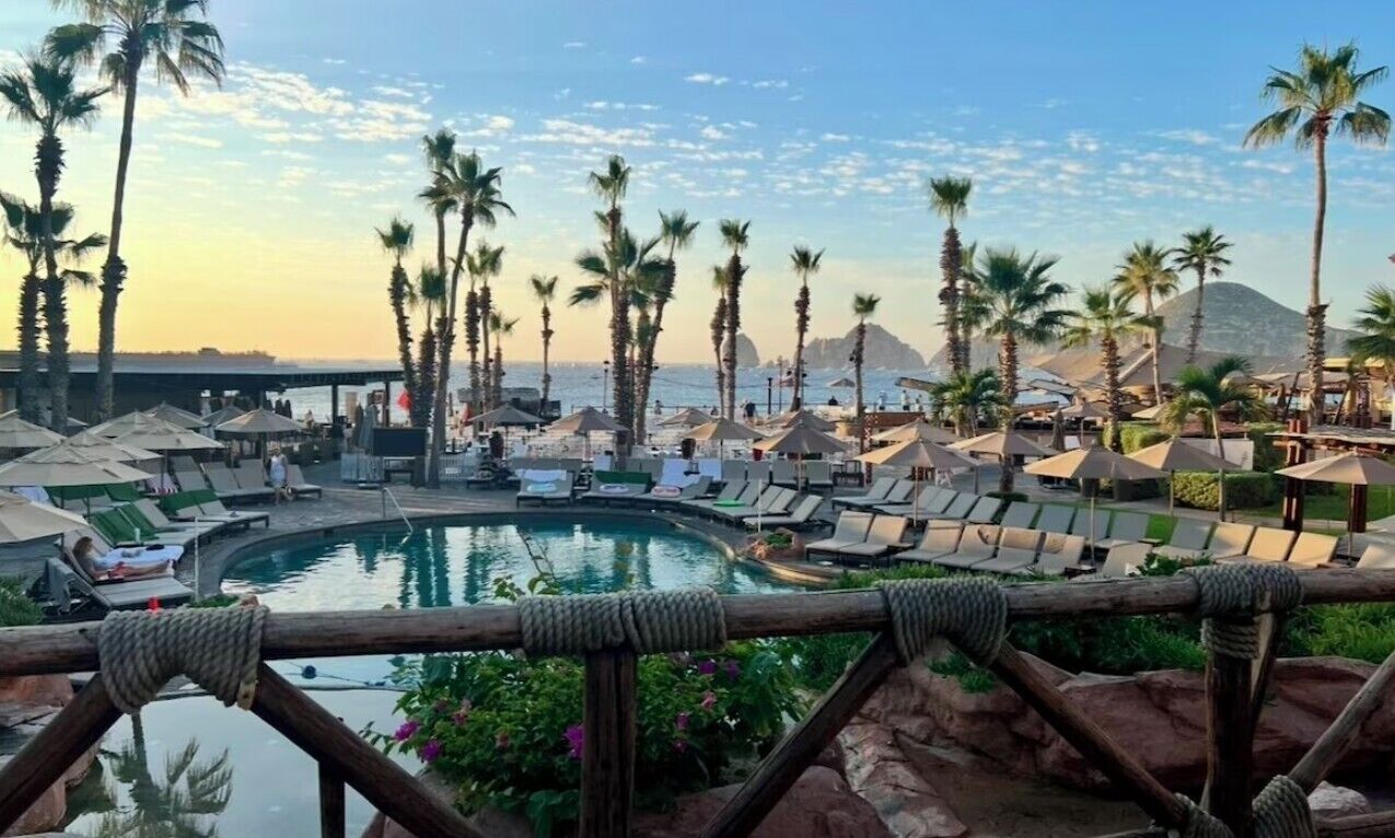 Zona de piscina en un resort con sillones de descanso y sombrillas, rodeada de palmeras y una montaña al fondo.