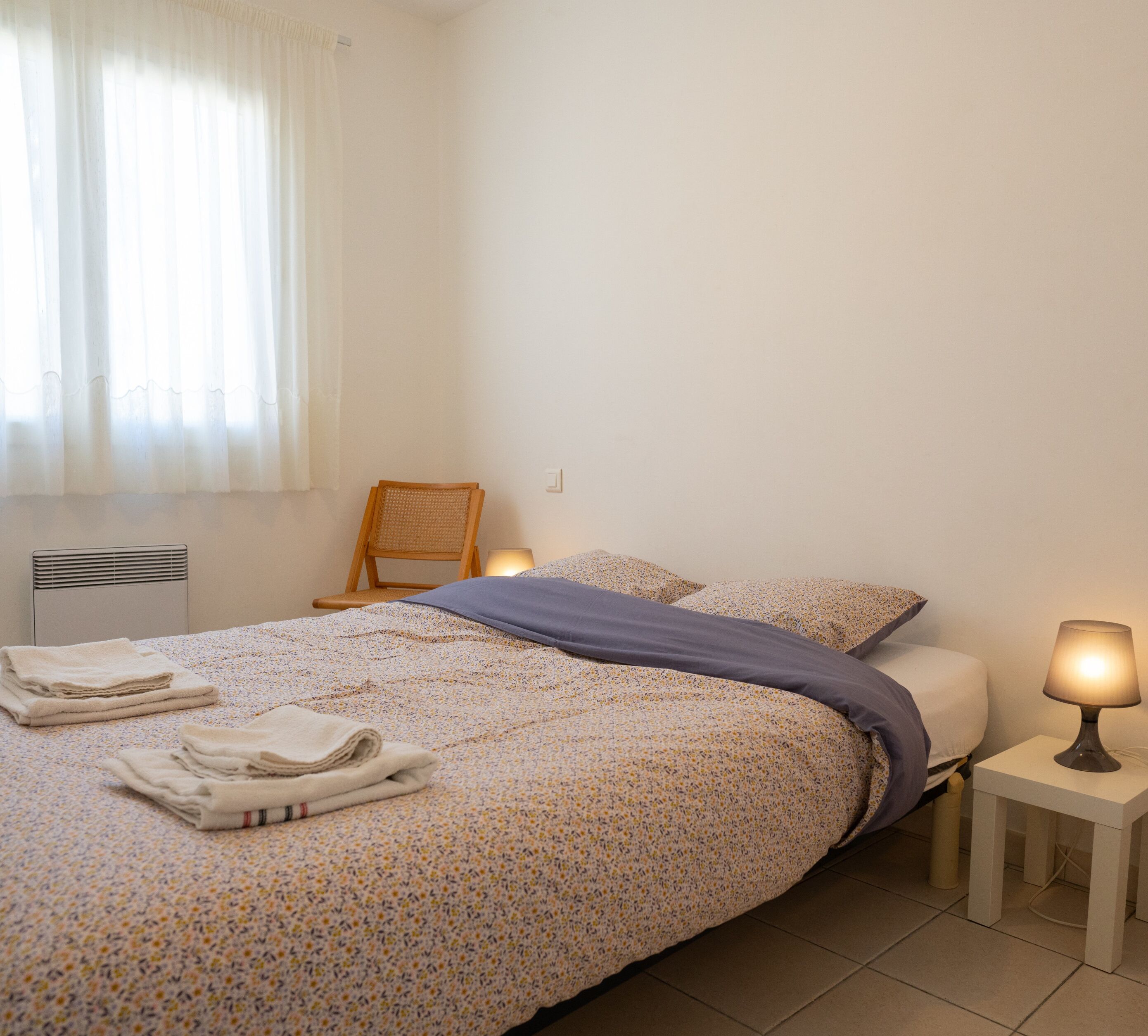 A neatly made bed with a floral-patterned bedspread and pillows, a chair, a bedside lamp, and a radiator.
