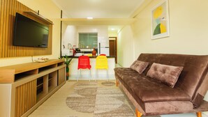 A living room with a brown sofa, a flat-screen TV mounted on a wooden panel, and a kitchen area with a refrigerator and a microwave.