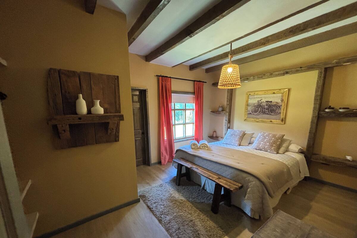 Quarto com cama, um banquinho, prateleira de madeira, quadro e luminária pendente.