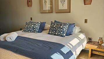 Ein Bett mit einer Decke, Kissen und einem Teppich. An den Wänden sind Leuchten angebracht und ein kleiner Tisch mit einer Vase steht daneben.