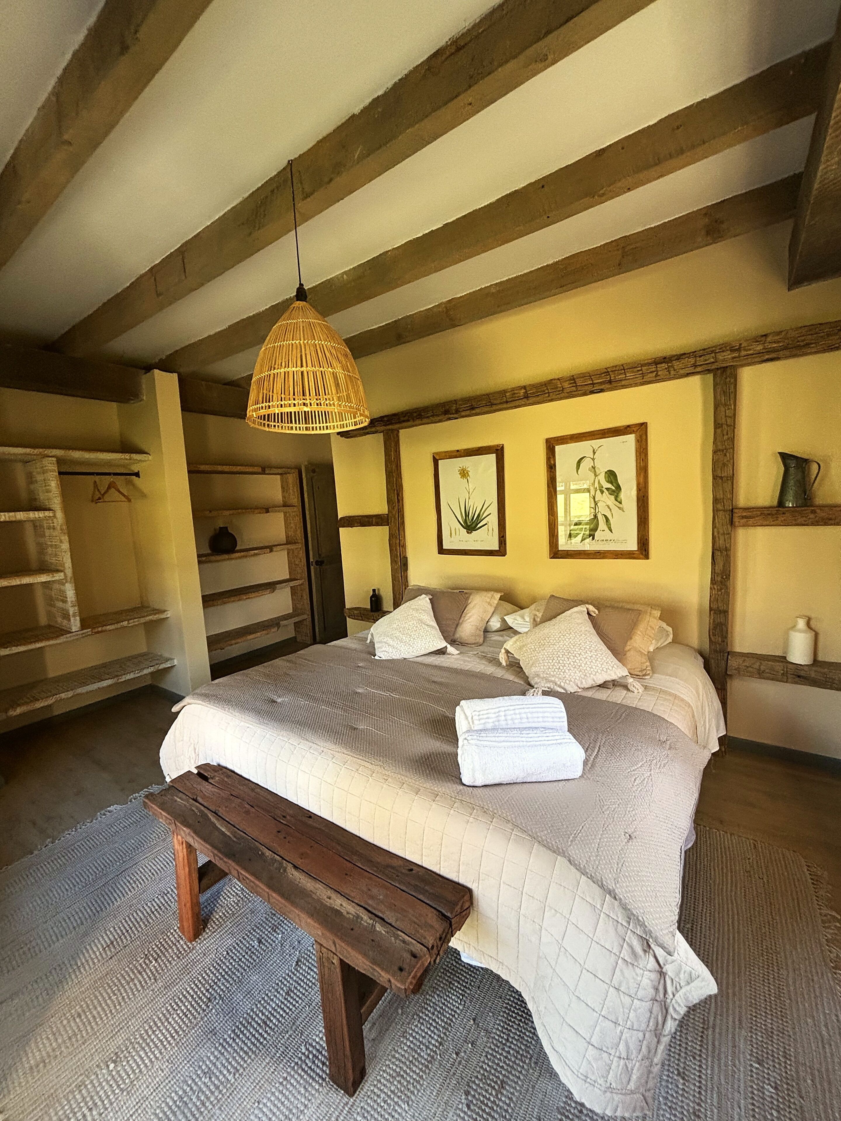A bedroom with a large bed, wooden beams, a hanging light fixture, and framed botanical prints on the wall.