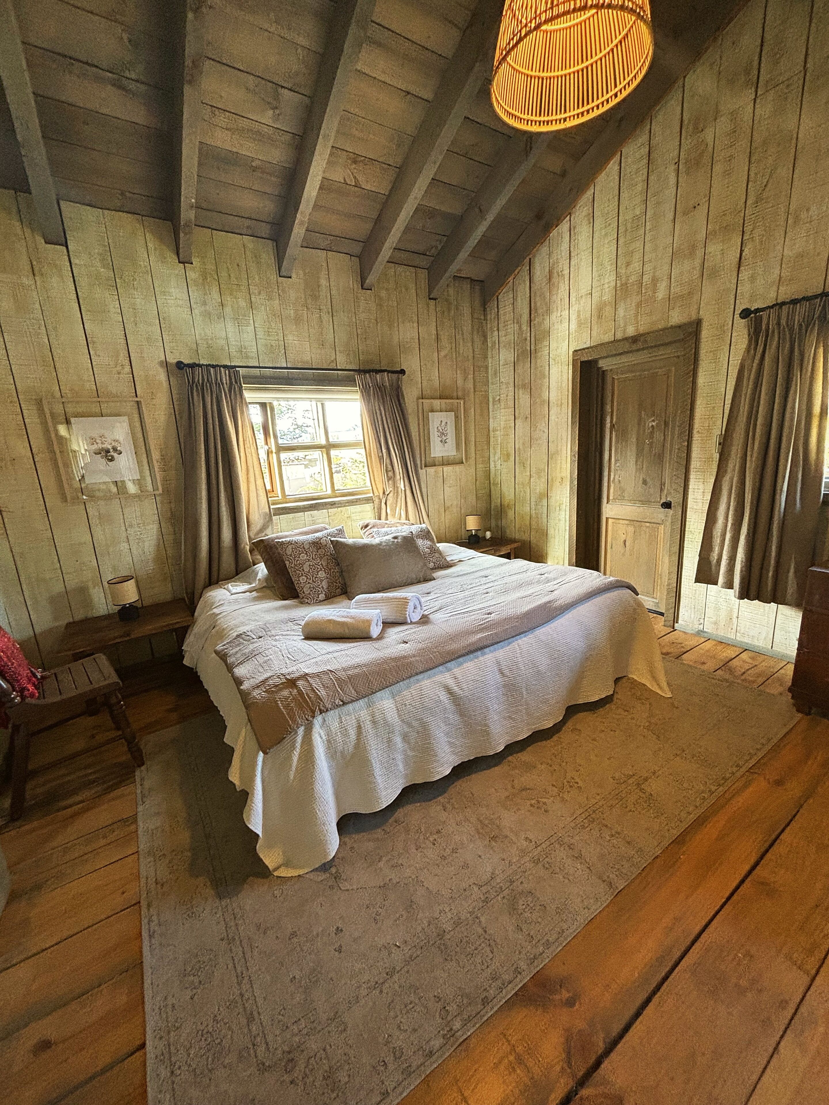 Una habitación de madera con una cama, una ventana, una puerta y una lámpara colgante.