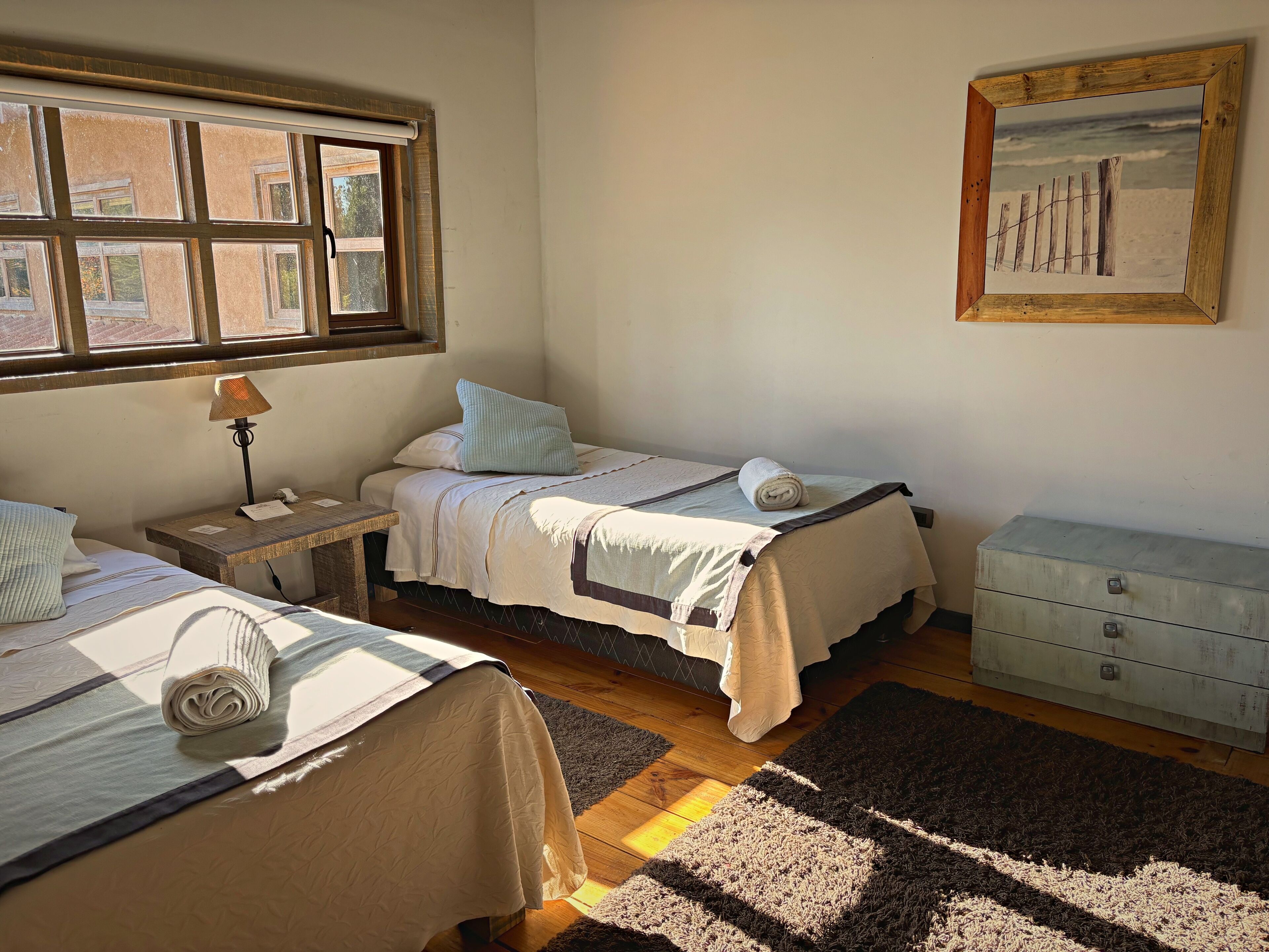 A bedroom with two beds, a nightstand, a dresser, a window with blinds, and a framed picture on the wall.