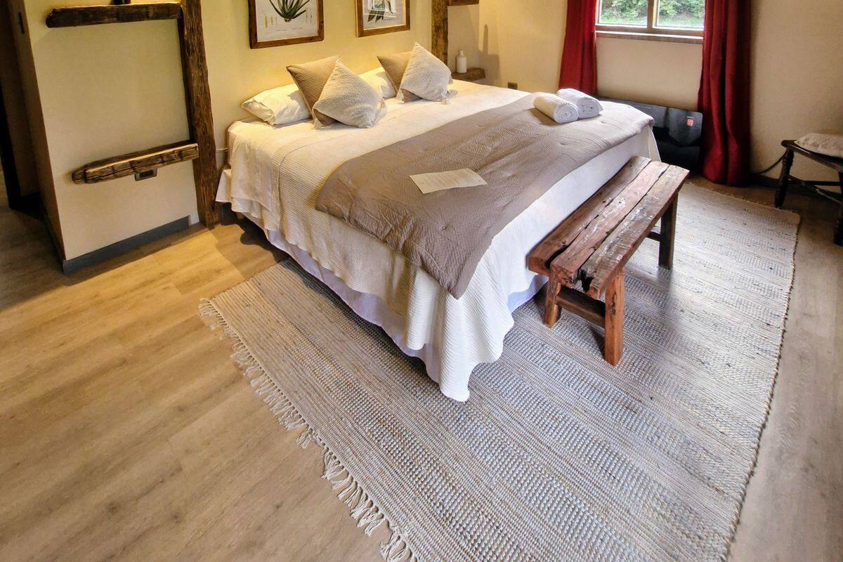 Quarto com cama grande, vigas de madeira, luminária pendente e impressões botânicas emolduradas na parede.