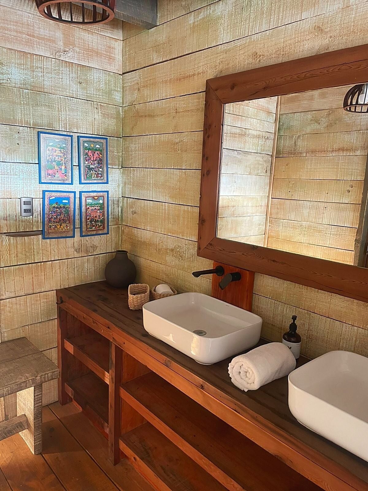 Une salle de bain en bois avec un meuble-lavabo en bois, deux vasques, un miroir et quatre tableaux encadrés accrochés au mur.