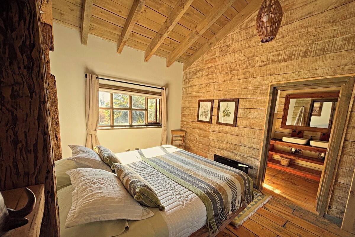 Quarto com teto de madeira, uma cama grande com várias almofadas, uma janela e um banheiro visível através de uma porta aberta.