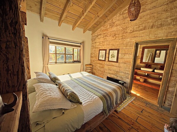 Une chambre avec un plafond en bois, un grand lit avec de nombreux oreillers, une fenêtre et une salle de bain visible à travers une porte ouverte.