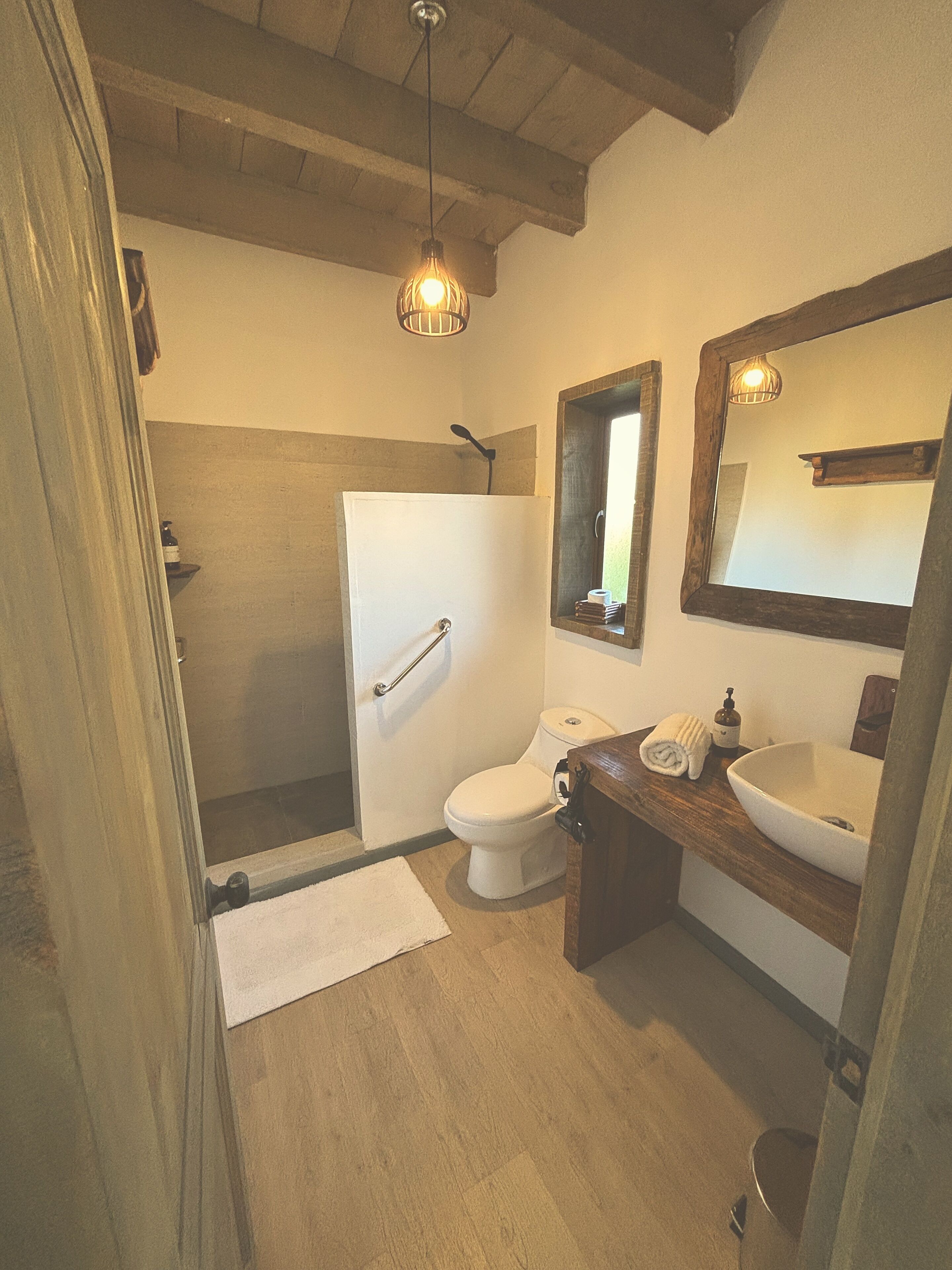 Un bany amb terra de fusta, un vàter blanc, un lavabo amb un mirall damunt i una zona de dutxa.