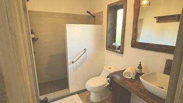 Un bany amb terra de fusta, un vàter blanc, un lavabo amb un mirall damunt i una zona de dutxa.