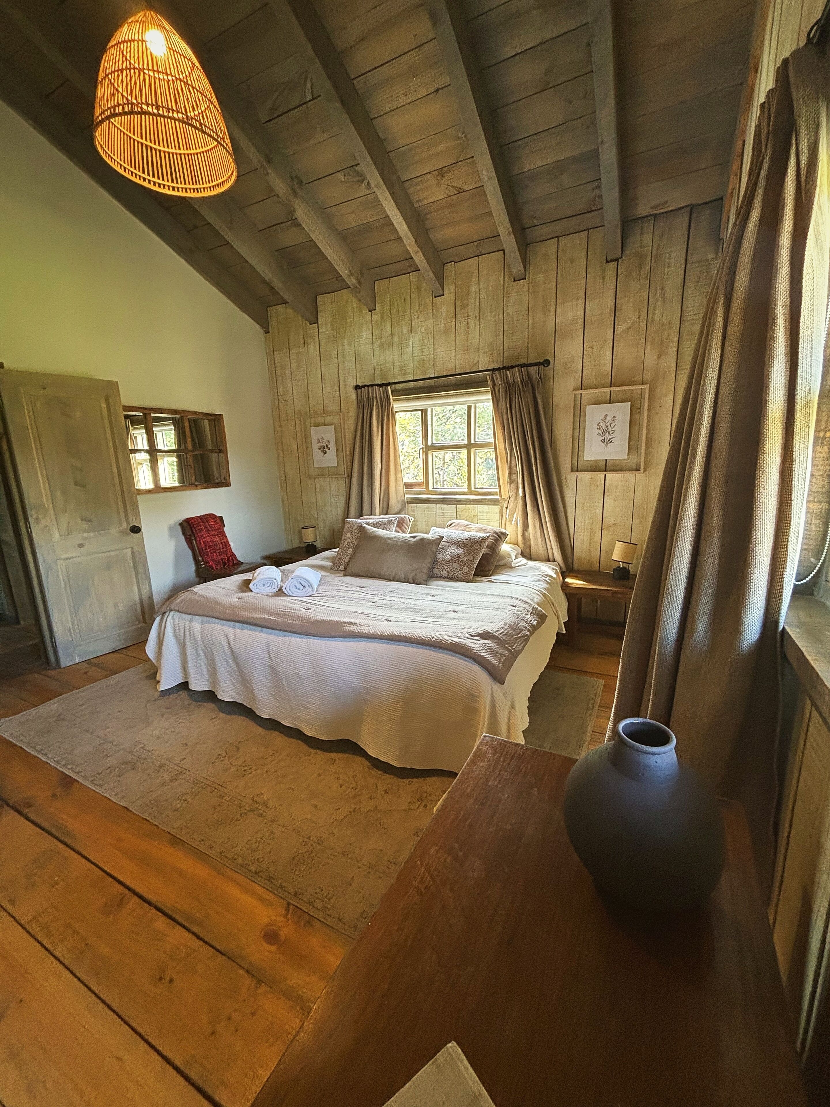 A bedroom with a wooden ceiling, a bed with multiple pillows, a window with curtains, and a hanging light fixture.