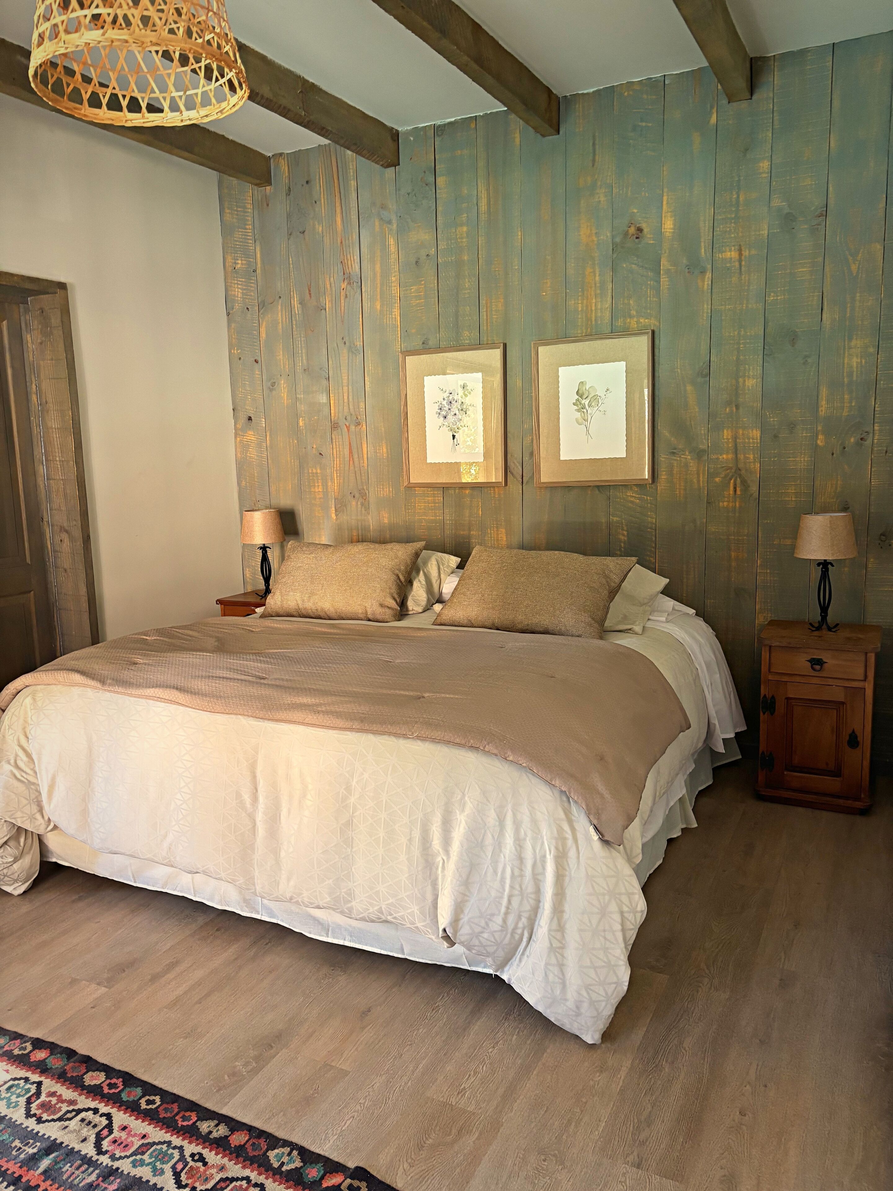A bedroom with a wooden wall, a bed with beige bedding, two framed pictures, and a nightstand with a lamp.
