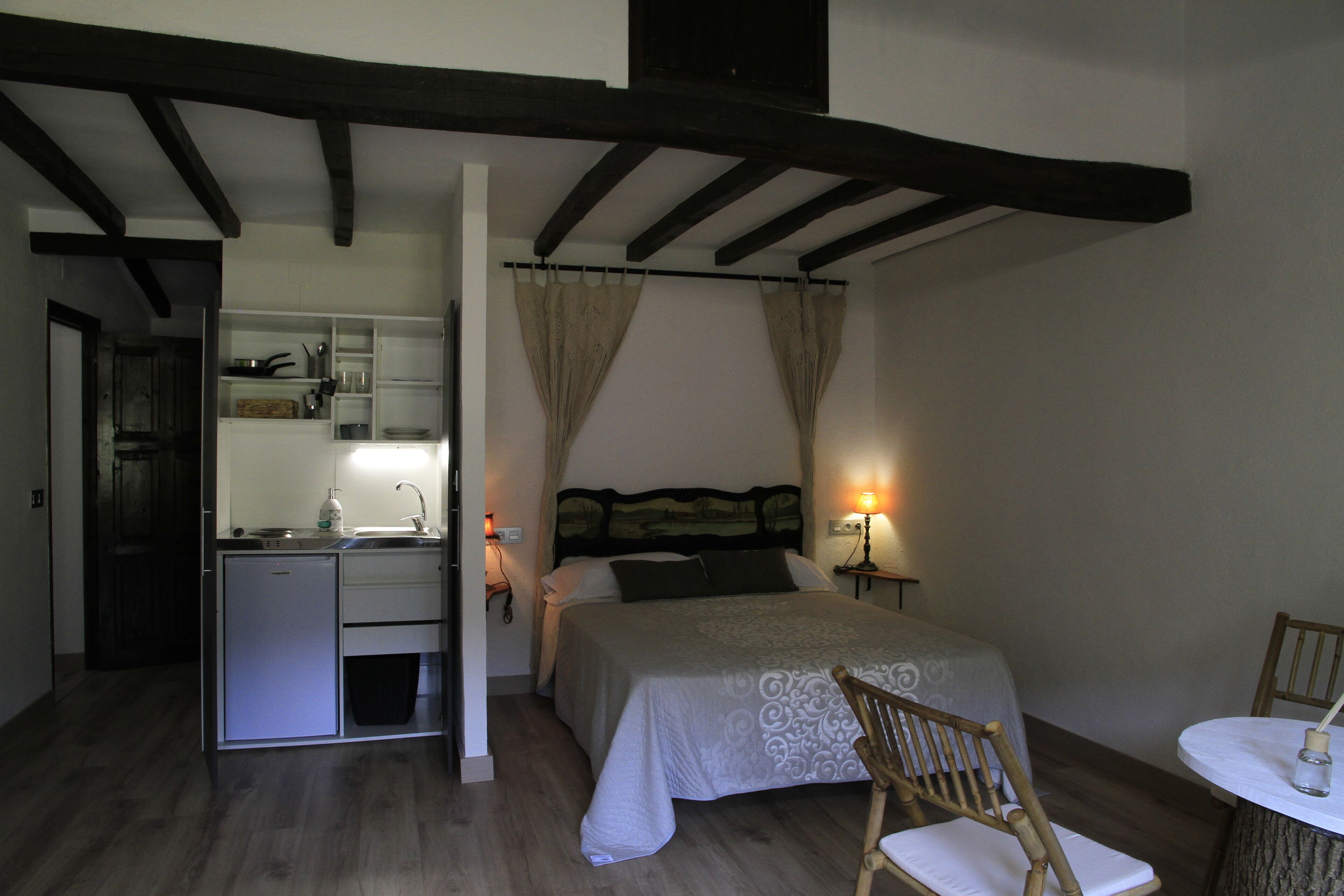 A compact bedroom with a bed, bedside table, chair, and a small kitchenette.