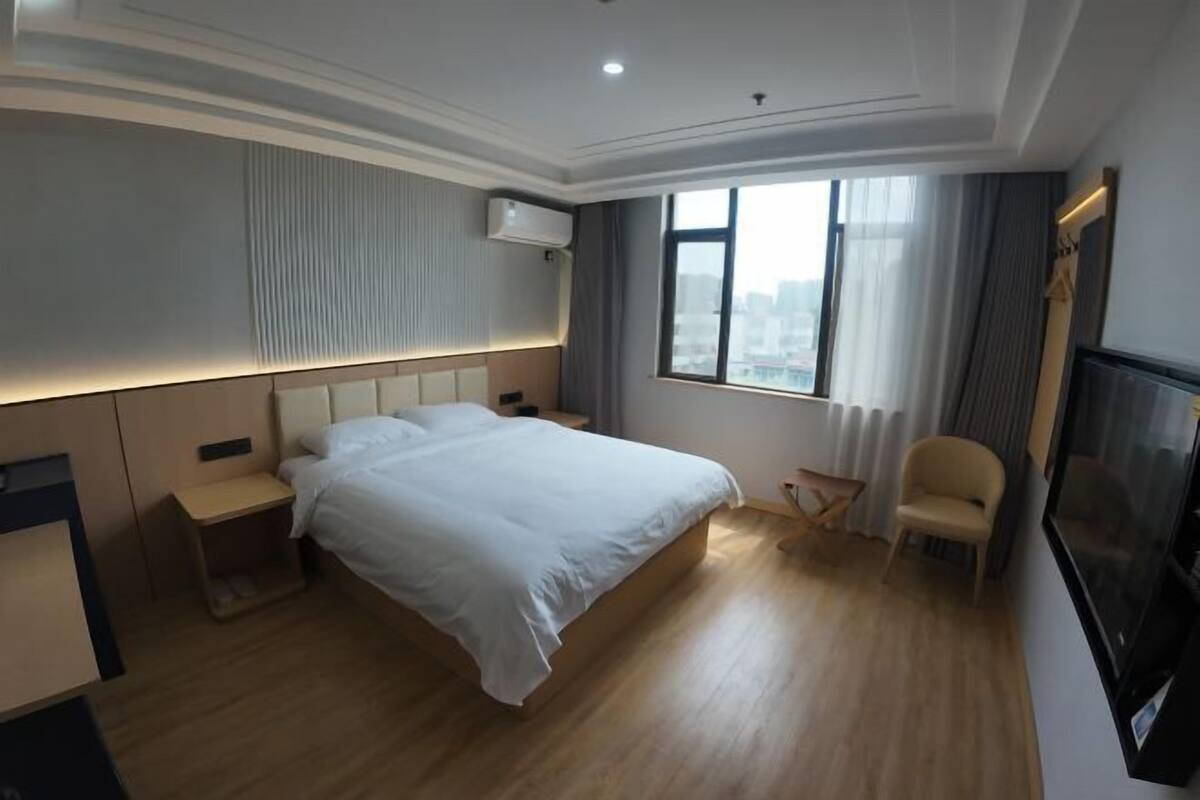 Ein Hotelzimmer mit einem großen Bett, Nachttischen, einem Sessel, einem Fernseher und einem Fenster mit Blick auf die Stadt.