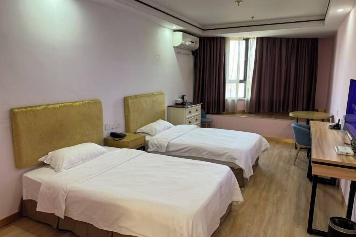 Ein Hotelzimmer mit zwei Einzelbetten, einem Schreibtisch, einem Stuhl, einem kleinen Tisch und einem Fenster mit Vorhängen.