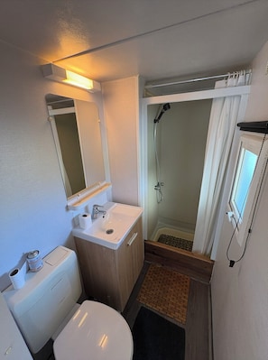A compact bathroom with a toilet, sink, mirror, shower, and a small window.