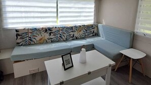 Un salon moderne avec un canapé d’angle, une petite table blanche et une fenêtre équipée de stores.