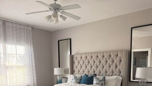 A bedroom with a tufted headboard, a white bedspread, two bedside lamps, a large mirror, and a ceiling fan.