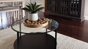 A modern living room with a glass coffee table, a potted plant, and a wooden tray with decorative items. A flat-screen TV is mounted on the wall, and a dark wooden cabinet is visible.