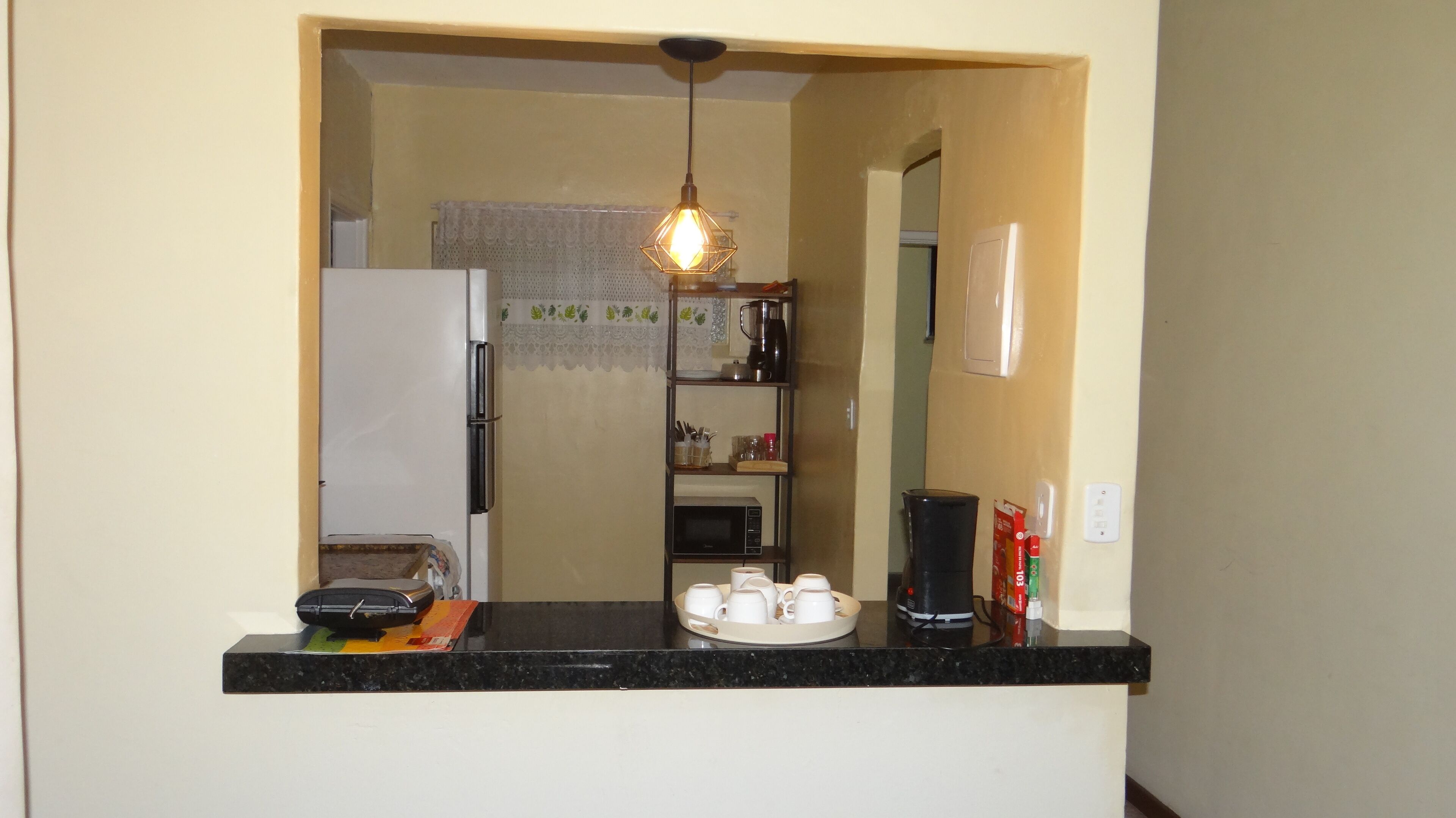 A kitchen with a refrigerator, microwave, coffee maker, and a shelf with cups and condiments.