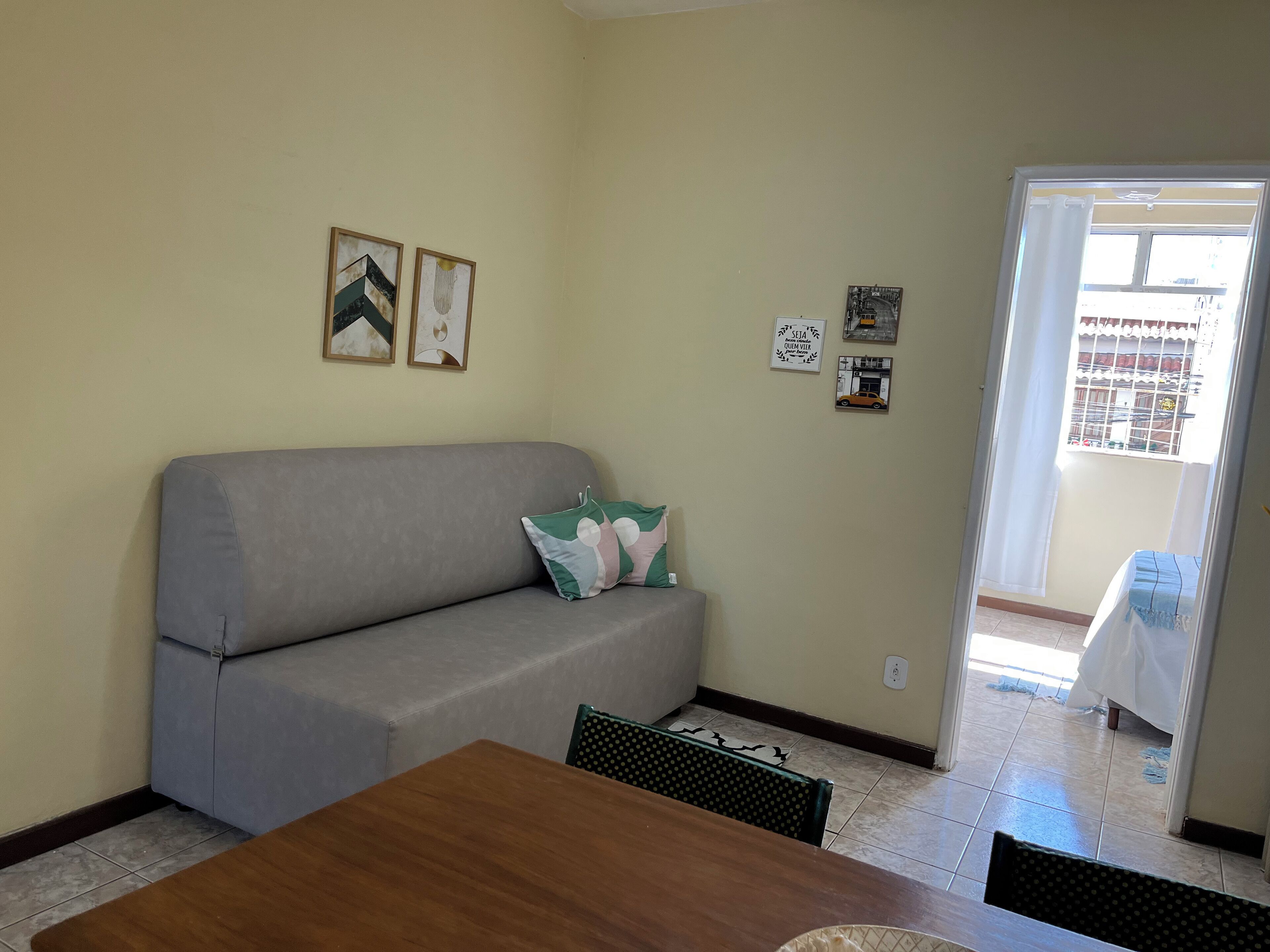 Ein Wohnzimmer mit grauem Sofa, einem Holzesstisch und einem gemusterten Sessel. An den Wänden sind Dekorationen angebracht und durch eine Tür ist ein Blick ins Freie möglich.