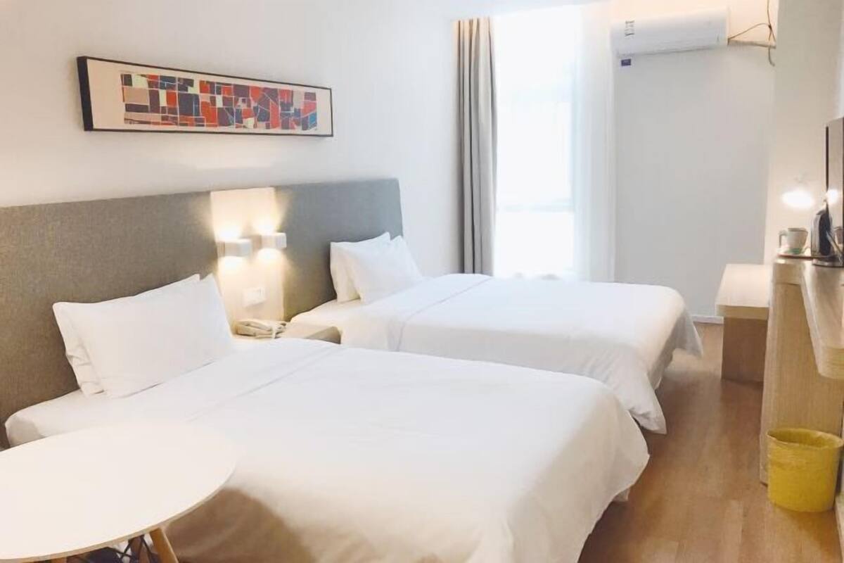 A hotel room with two beds, a small table, a bedside lamp, a painting on the wall, and an air conditioning unit.