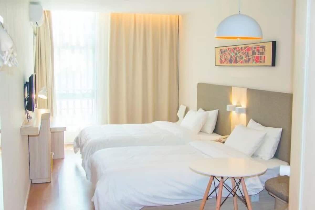 A hotel room with two beds, a small table, a desk, and a large window with curtains.