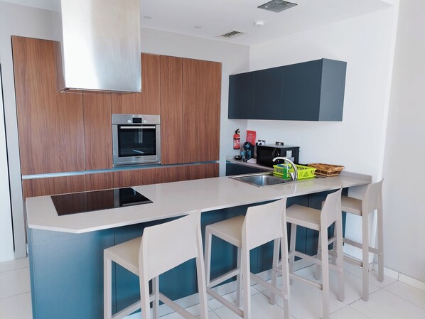 A modern kitchen with a built-in oven, a coffee maker, and a sink. There are three white bar stools and a large island with a digital tablet.