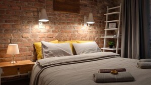 Ein Schlafzimmer mit Backsteinwand, einem Bett mit weißen und gelben Kissen, einem Nachttisch mit Lampe sowie einem Regal mit Pflanze.