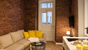 Ein gemütliches Wohnzimmer mit Backsteinwand, einer weißen Tür, einem Sofa mit gelben Kissen, einem kleinen runden Tisch und einem flachbildfernseher an der Wand.