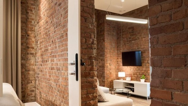Ein modernes Zimmer mit freiliegenden Backsteinwänden, einem Wand-Fernseher, einem weißen Sofa und einem kleinen Tisch mit einer Lampe.
