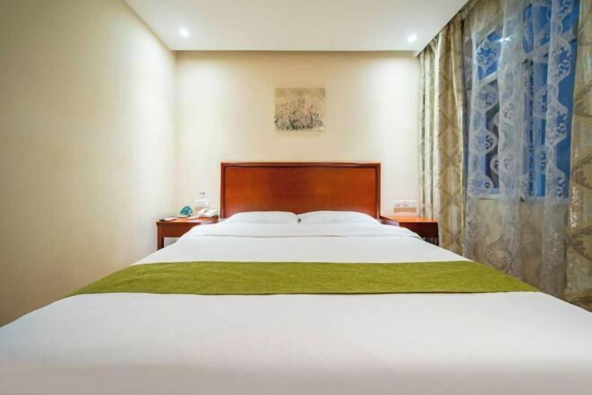 A hotel room with a large bed, a wooden headboard, a nightstand, a wall-mounted lamp, and a decorative painting.