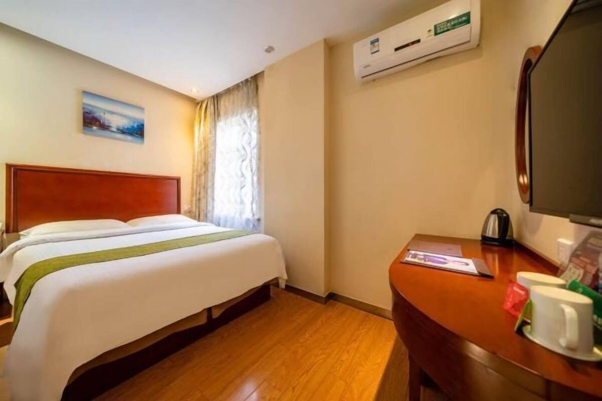A hotel room with a bed, a desk, a chair, an air conditioning unit, and a painting on the wall.