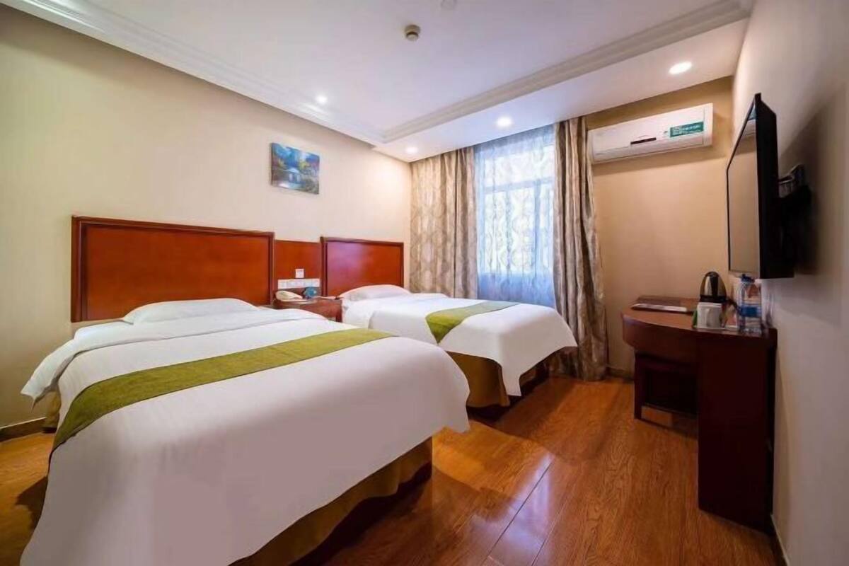 A hotel room with two beds, a wooden floor, a desk with a chair, a TV, and an air conditioning unit.