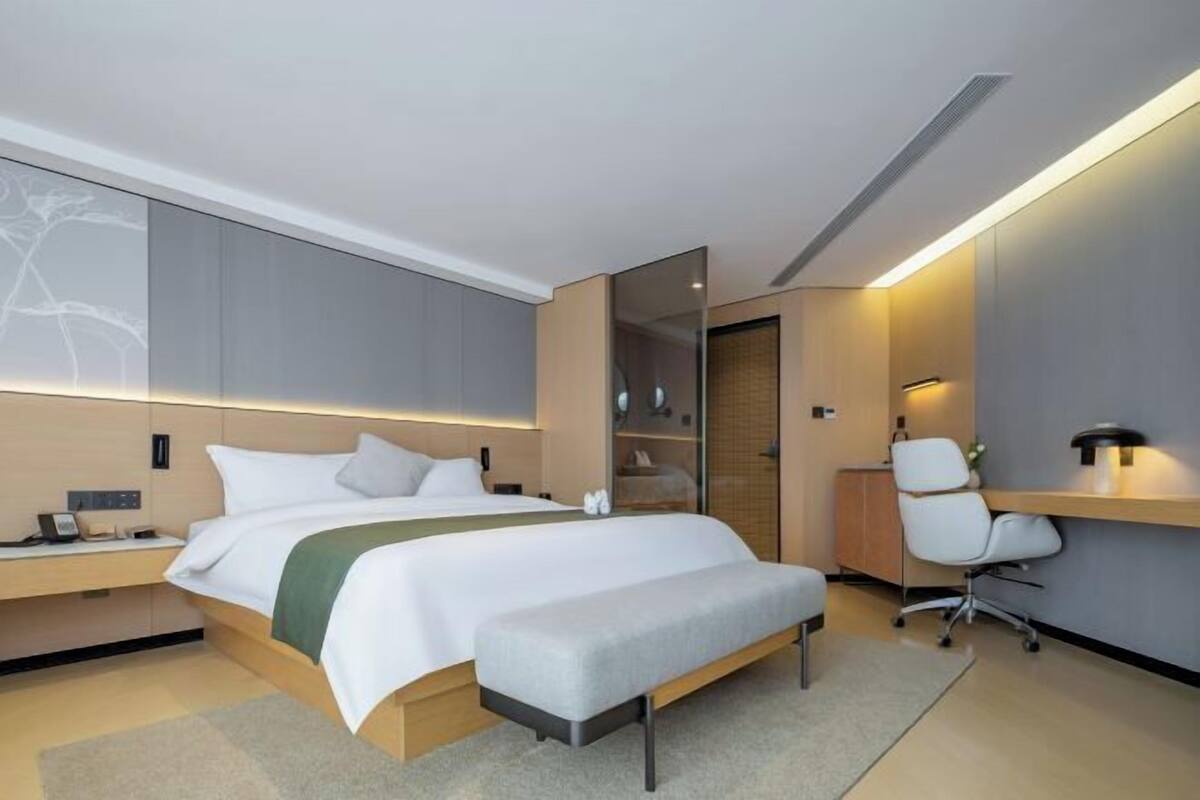 A modern hotel room with a bed, bedside table, desk, chair, and a wardrobe.