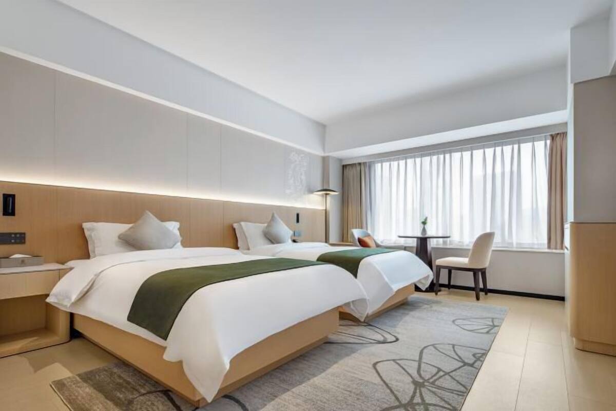A hotel room with two beds, a desk, a chair, and a large window with curtains.