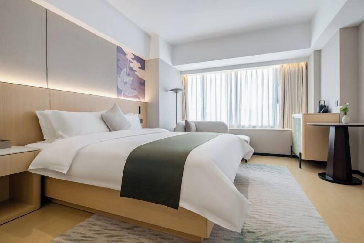A modern hotel room with a large bed, bedside tables, a desk, and a window with curtains.