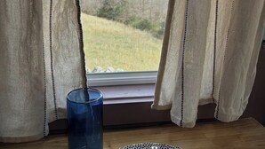 Una ventana con cortinas translúcidas, un vaso de vidrio azul y un plato blanco con un diseño azul sobre una mesa de madera.