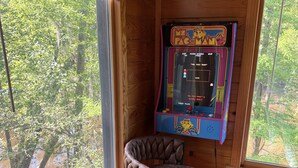 Et trægulvsrum med et gammelt Pac-Man arkadespil, en polstret stol og store vinduer med udsigt over et skovområde.